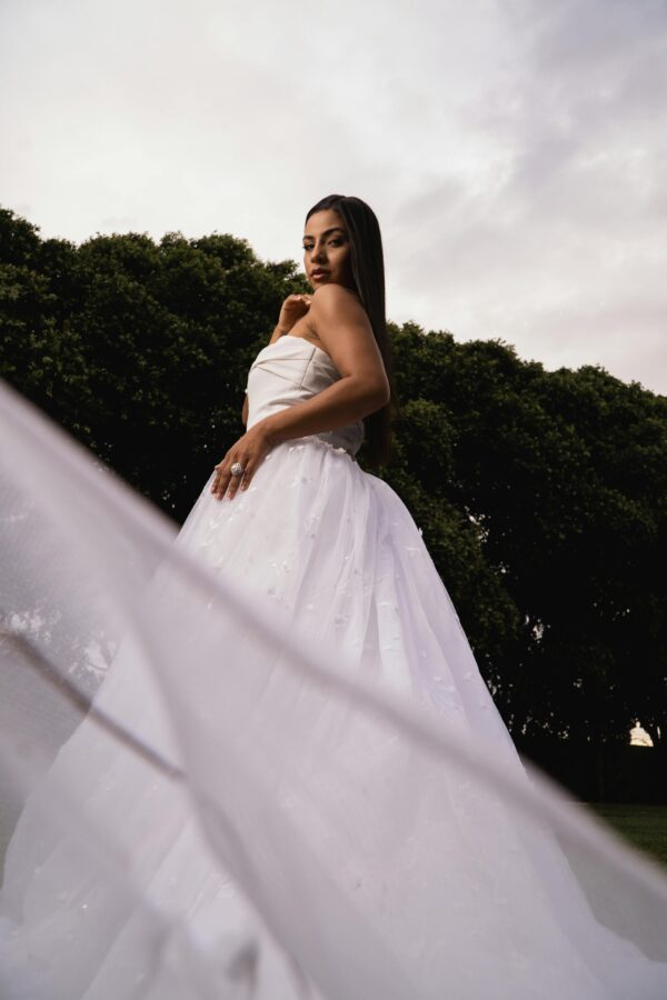 pexels-photo-32498326-32498326 Beautiful bride posing in a white gown, showcasing elegance and grace against lush green trees.
